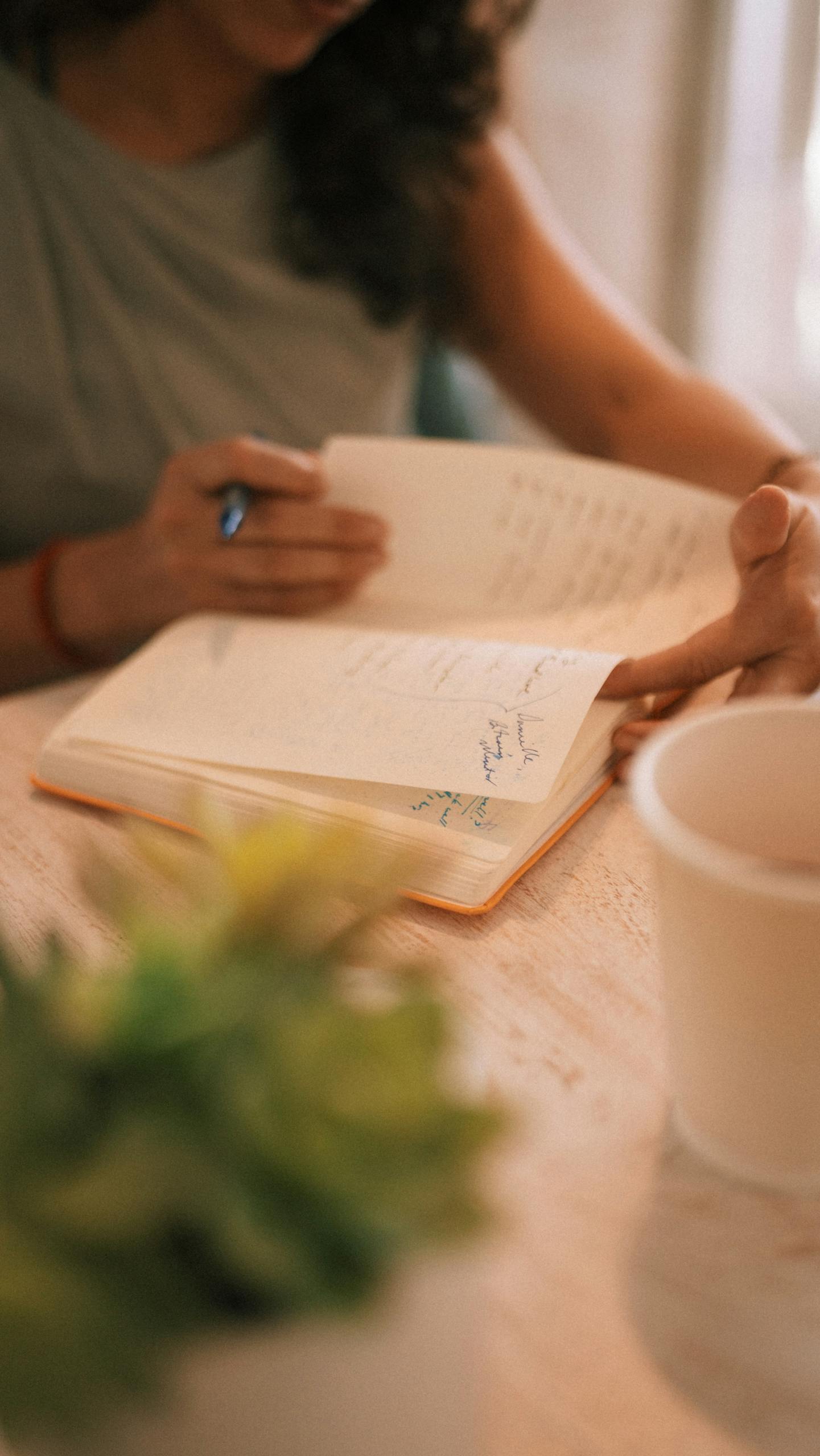 Close-up of a person writing in a notebook with a coffee cup nearby, warm setting.